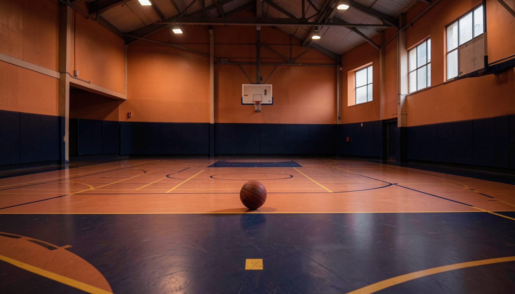 A wide indoor gymnasium bathed in warm terracotta and deep navy light, with a lone basketball resting near center court, evoking focus and quiet determination