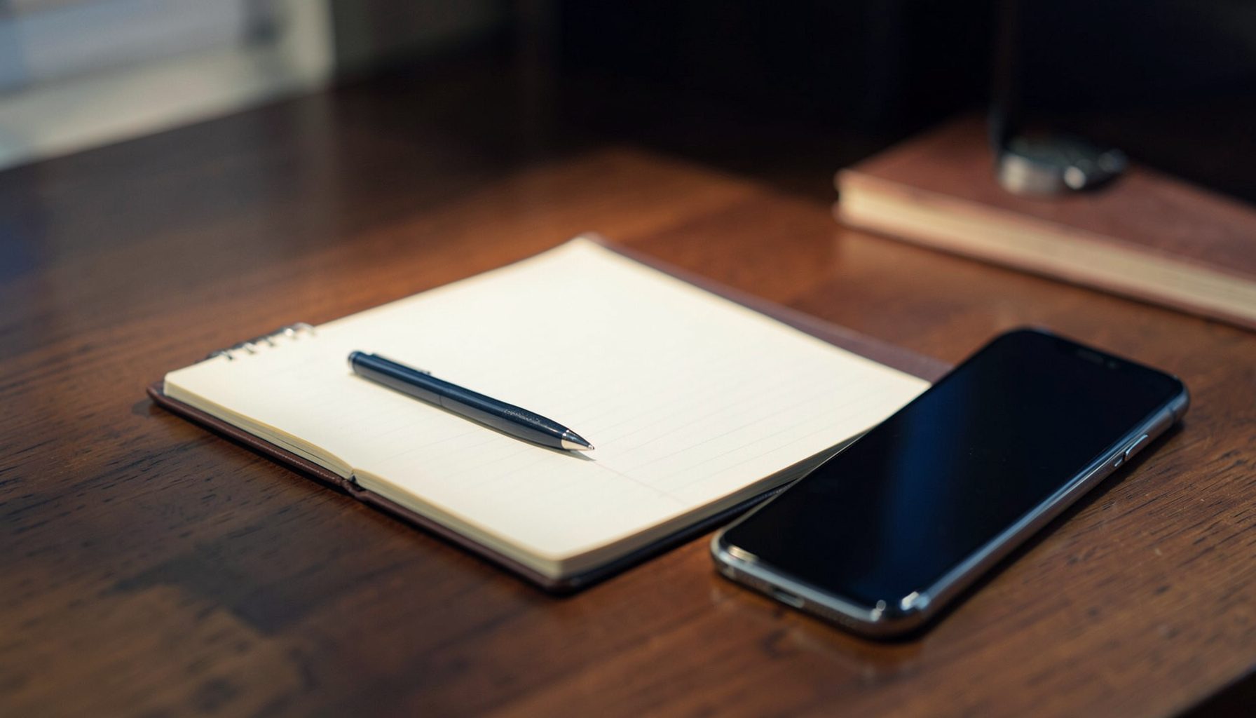 A softly lit desk scene with a notebook, pen, and a smartphone resting on a warm wooden surface, deep navy and cream tones